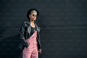 Giovane donna moderna alla moda che indossa occhiali da sole, cappello nero e giacca, in posa sul muro nero della città. Un sacco di spazio per la copia.