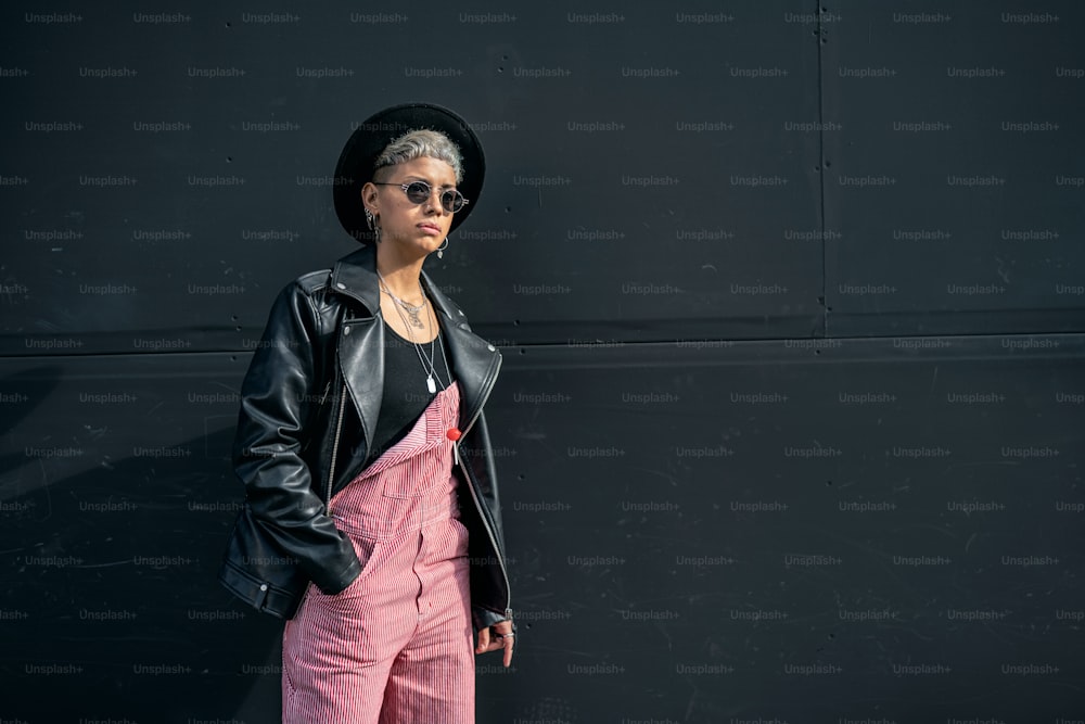 Giovane donna moderna alla moda che indossa occhiali da sole, cappello nero e giacca, in posa sul muro nero della città. Un sacco di spazio per la copia.