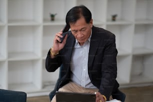 Asian senior manager and businessman in black suit relaxing in social media online and talking, calling with businesspeople on smartphone at workspace in office.