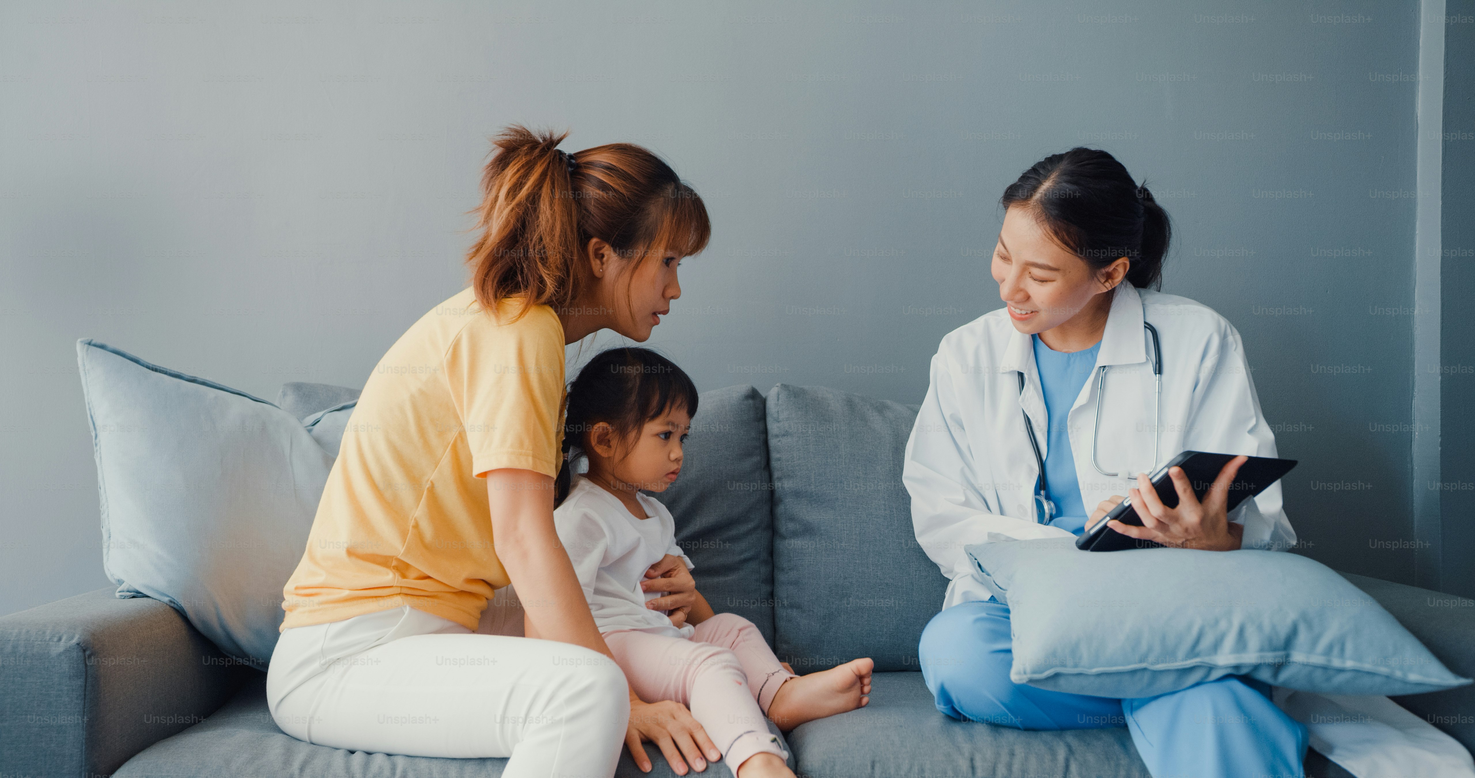 Dokter anak perempuan Asia muda dan pasien gadis kecil menggunakan tablet digital berbagi kabar tes kesehatan yang baik dengan ibu yang bahagia duduk di sofa di rumah.