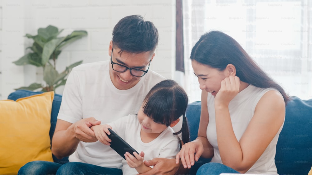 Felice famiglia asiatica papà, mamma e figlia che giocano a un gioco divertente online su smartphone seduto divano nella stanza di casa.