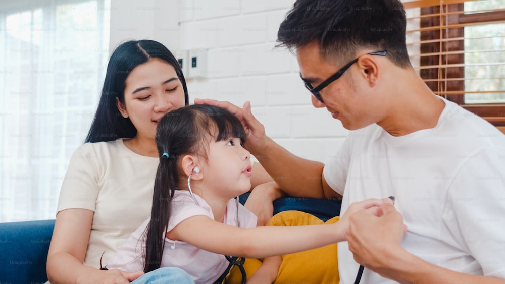 Happy cheerful Asian family dad, mom and daughter playing funny game as doctor having fun on sofa at home.