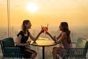 Belle amie asiatique se rencontrant et buvant un cocktail ensemble dans un restaurant sur le toit d’un gratte-ciel dans la métropole au coucher du soleil d’été. Une amie profite d’une activité de plein air dans la ville la nuit