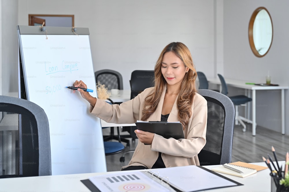 Confident businesswoman holding documents and writing business plan on whiteboard.