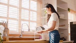 Atractiva joven asiática lavando platos mientras limpia en la cocina de la casa.