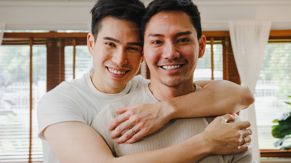 Ritratto Giovane Asia Coppia gay che si sente felice sorridente a casa. Gli uomini LGBTQ asiatici si rilassano con un sorriso a denti stretti guardando la telecamera mentre si abbracciano nel soggiorno di casa.