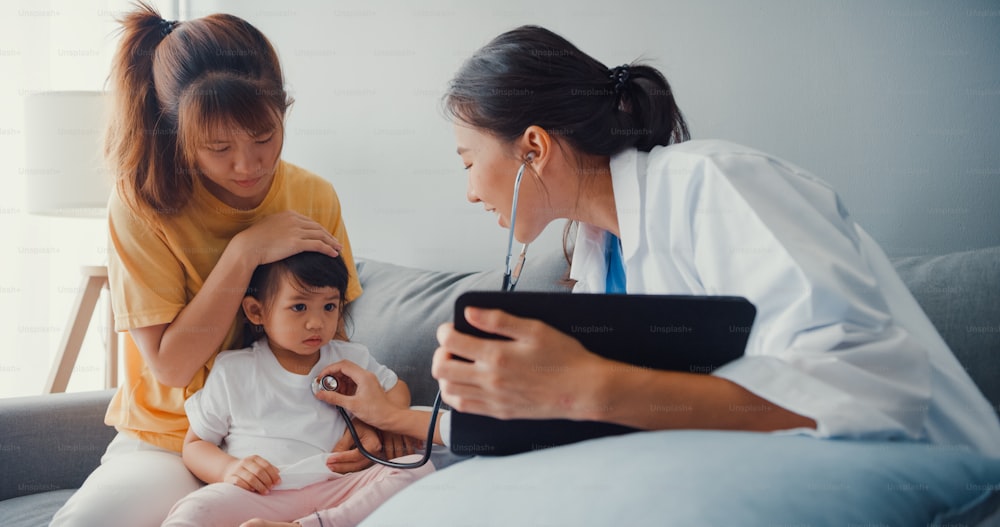 Young Asia female pediatrician hold stethoscope exam little girl patient visit doctor with mother sit on couch in living room at house.