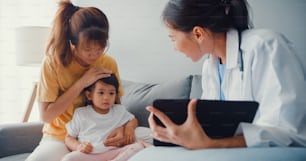 Jeune médecin pédiatre asiatique et petite fille patiente utilisant une tablette numérique partageant de bonnes nouvelles sur les tests de santé avec une maman heureuse assise sur le canapé dans la maison.