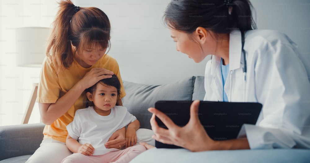 Junge asiatische Kinderärztin und kleine Patientin mit digitalem Tablet teilen gute Gesundheitstest-Nachrichten mit glücklicher Mutter sitzen auf der Couch im Haus.