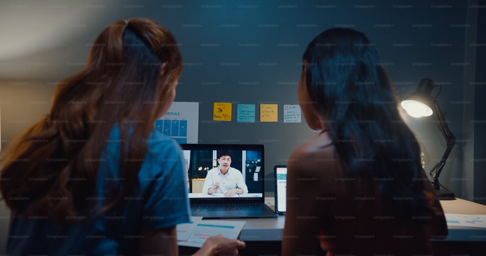 Young student Asia ladies watching lesson online and studying with roommate site on table in living room from home at night.