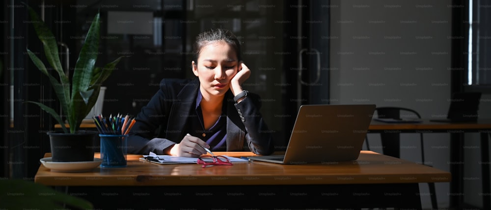 Jovem empresária cansada sentada em sua mesa de escritório com laptop.