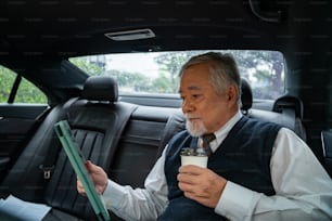 Confidence senior businessman in suit sitting on car backseat drinking hot coffee while reading business plan on digital tablet. Elderly executive CEO working in driving automobile going to office