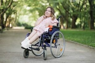 Porträt einer angenehmen jungen Frau mit spinaler Muskelatrophie, die vor der Kamera im grünen Sommerpark lächelt. Weibliche Person, die Rollstuhl benutzt. Konzept von Menschen mit Behinderung.