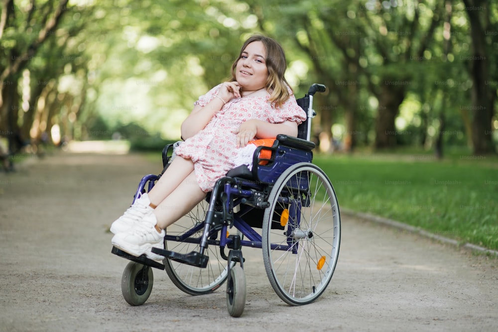 Portrait of pleasant young woman with spinal muscular atrophy smiling on camera among green summer park. Female person who using wheelchair. Concept of people with disability.