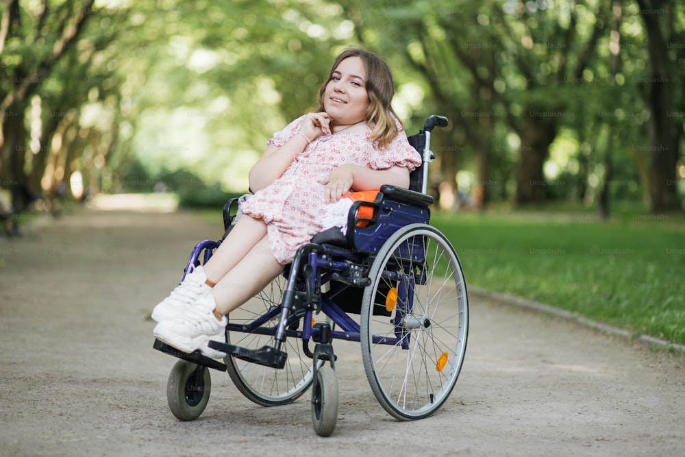 Retrato de una mujer joven agradable con atrofia muscular espinal sonriendo en la cámara entre el parque verde del verano. Persona femenina que utiliza silla de ruedas. Concepto de personas con discapacidad.