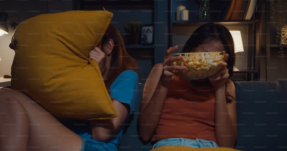 Attractive Asia couple ladies feel shock and fear moment eat popcorn watch horror online movie entertainment on sofa  in living room at home dark night.