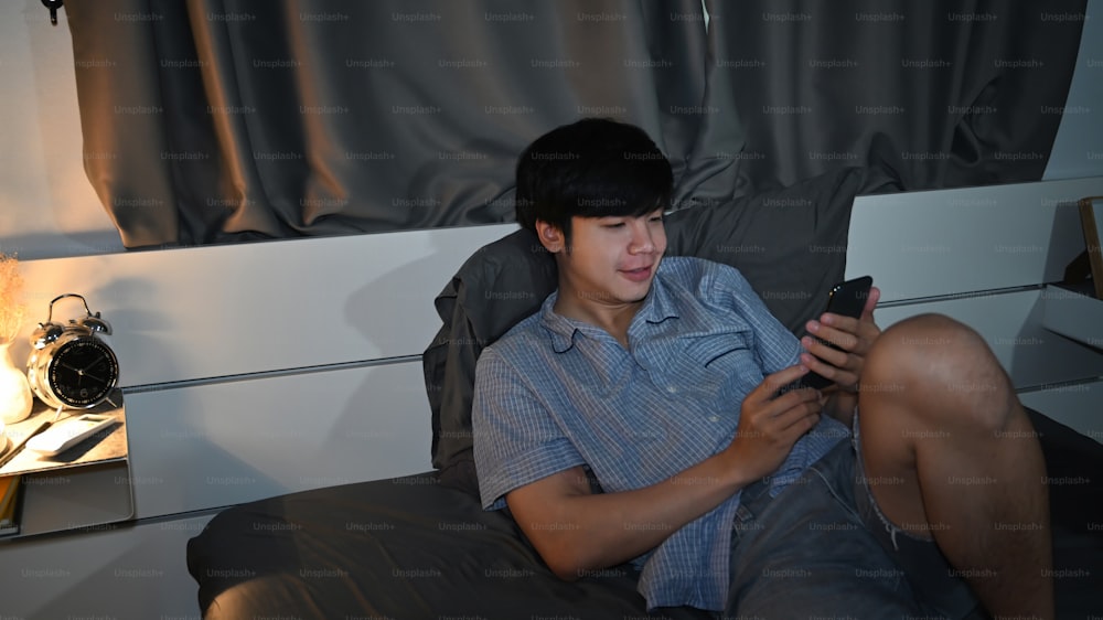 Hombre asiático feliz acostado en la cama y revisando las redes sociales con un teléfono inteligente.