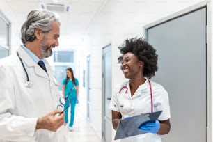 Surgeon and Female Doctor Walk Through Hospital Hallway, They Consult while Talking about Patient's Health looking at clipboard. Modern Bright Hospital with Professional Staff.