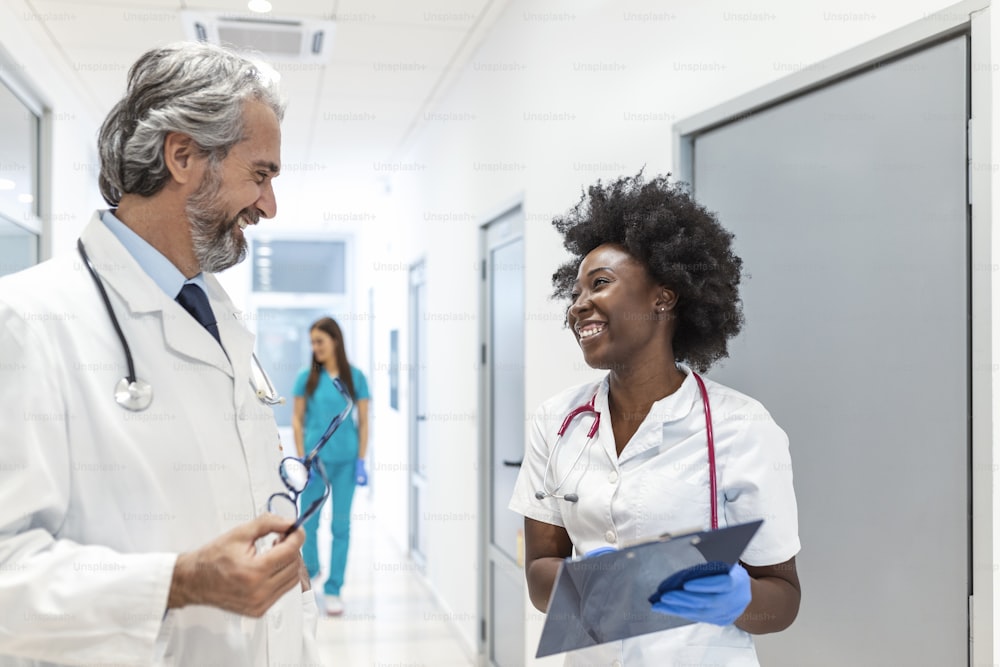 Un chirurgien et une femme médecin traversent le couloir de l’hôpital, ils consultent tout en parlant de la santé du patient en regardant le presse-papiers. Hôpital moderne et lumineux avec un personnel professionnel.