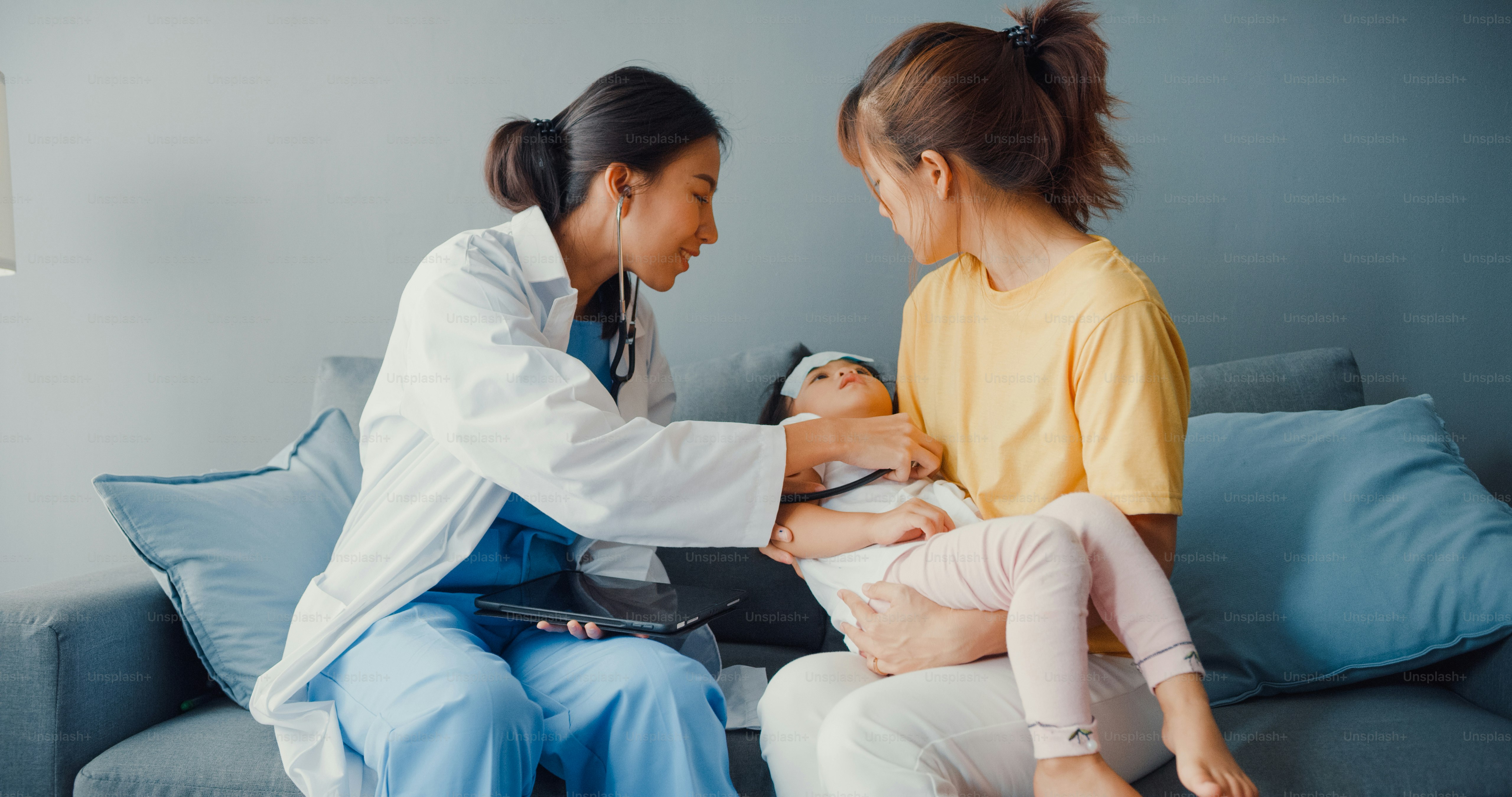 Young Asia female pediatrician hold stethoscope exam little girl patient visit doctor with mother sit on couch in living room at house.