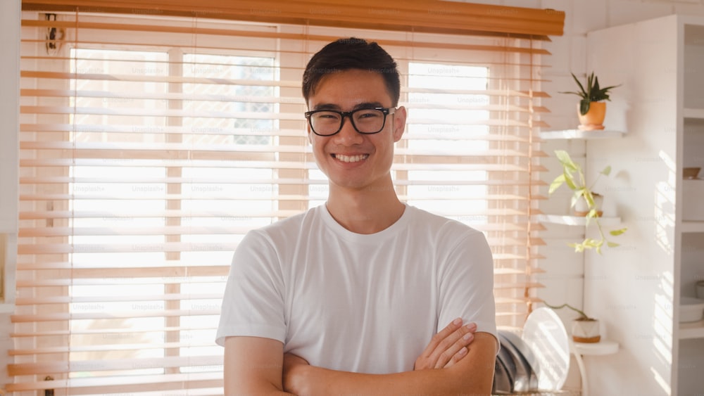 Glücklicher junger asiatischer Mann, der glücklich lächelt, die Arme verschränkt und in die Kamera schaut, während er sich in der Küche zu Hause entspannt.