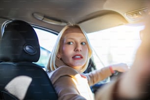 Woman in car indoor turning around looking at passengers in back seat idea taxi driver. Concept of exam Vehicle. Back view of an attractive young woman looking over her shoulder while driving a car.
