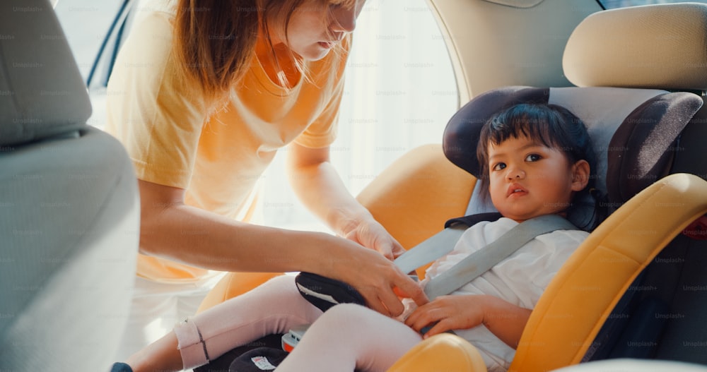 Happy Asia family mom fastening seat belt of child car seat before go for ride with toddler girl preparing for trip in car. Safety driving concept.