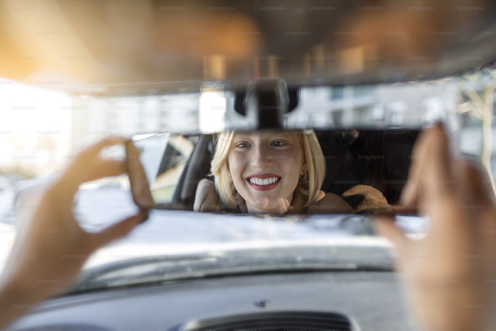 Mujer ajustando a mano el espejo retrovisor de su coche. Conductora joven feliz que busca ajustar el espejo retrovisor del automóvil, asegurándose de que la línea esté libre, la visibilidad es buena