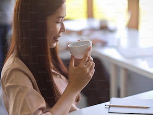 A beautiful, relaxed businesswoman is sipping hot coffee or tea at her office workplace. Feeling clam and peaceful  close-up