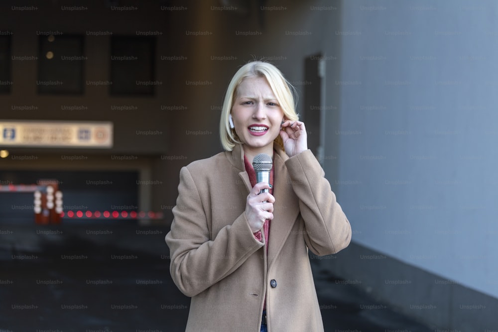 Retrato recortado de una reportera profesional en el trabajo. Mujer joven parada en la calle con un micrófono en la mano y sonriendo a la cámara. Disparo horizontal. Enfoque selectivo en la mujer