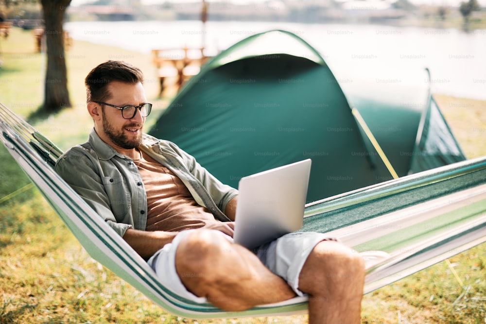 Mid adult man relaxing in hammock while working on laptop during camping day in nature.
