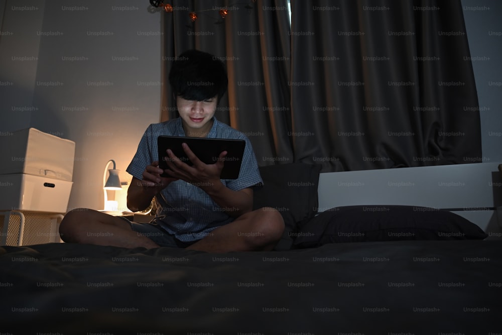 Young asian man using digital tablet while sitting on comfortable bed at night time.