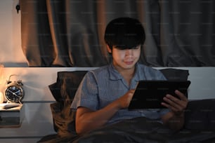 Young man using digital tablet while sitting on the bed at night time.