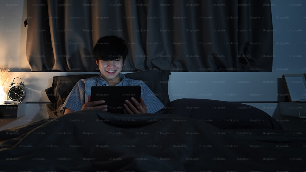 Jovem sentado na cama e assistindo a filmes ou navegando na internet com tablet digital à noite.