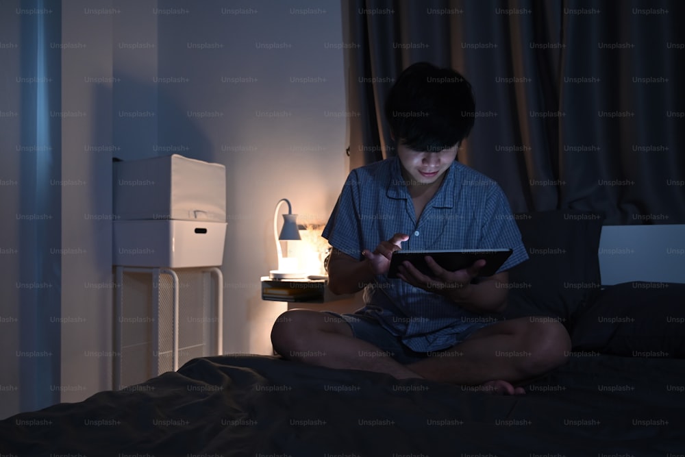 Jeune homme utilisant une tablette numérique travaillant tard le soir sur son lit.