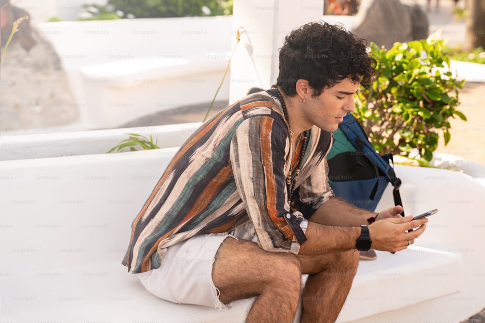 Cheerful young spanish man sitting outdoor and using mobile phone, scrolling through social media. Traveler guy talking online at telephone. Modern concept of travel and web technology. Real people lifestyle.
