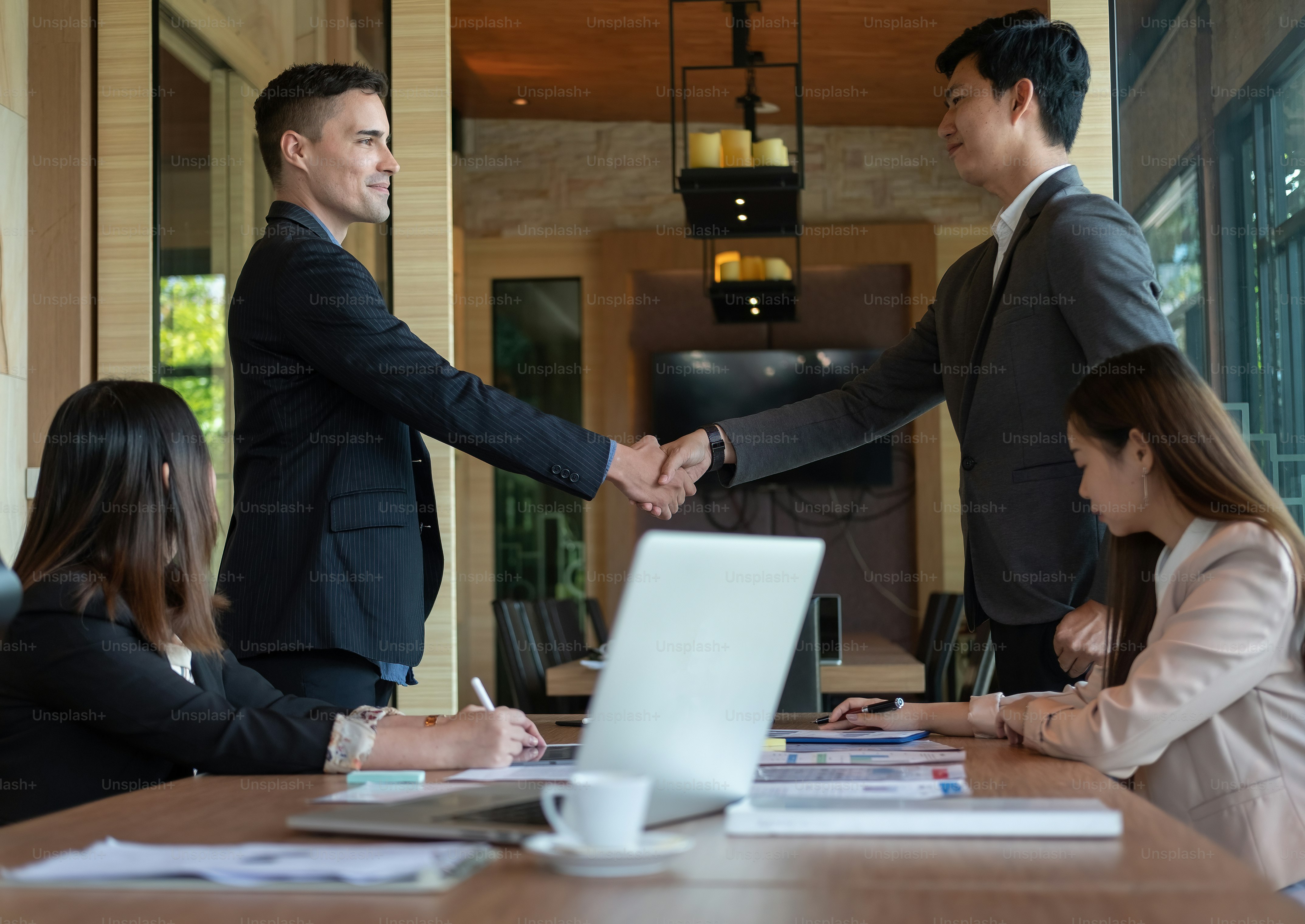 Two men in suits shaking hands across a conference table with two women