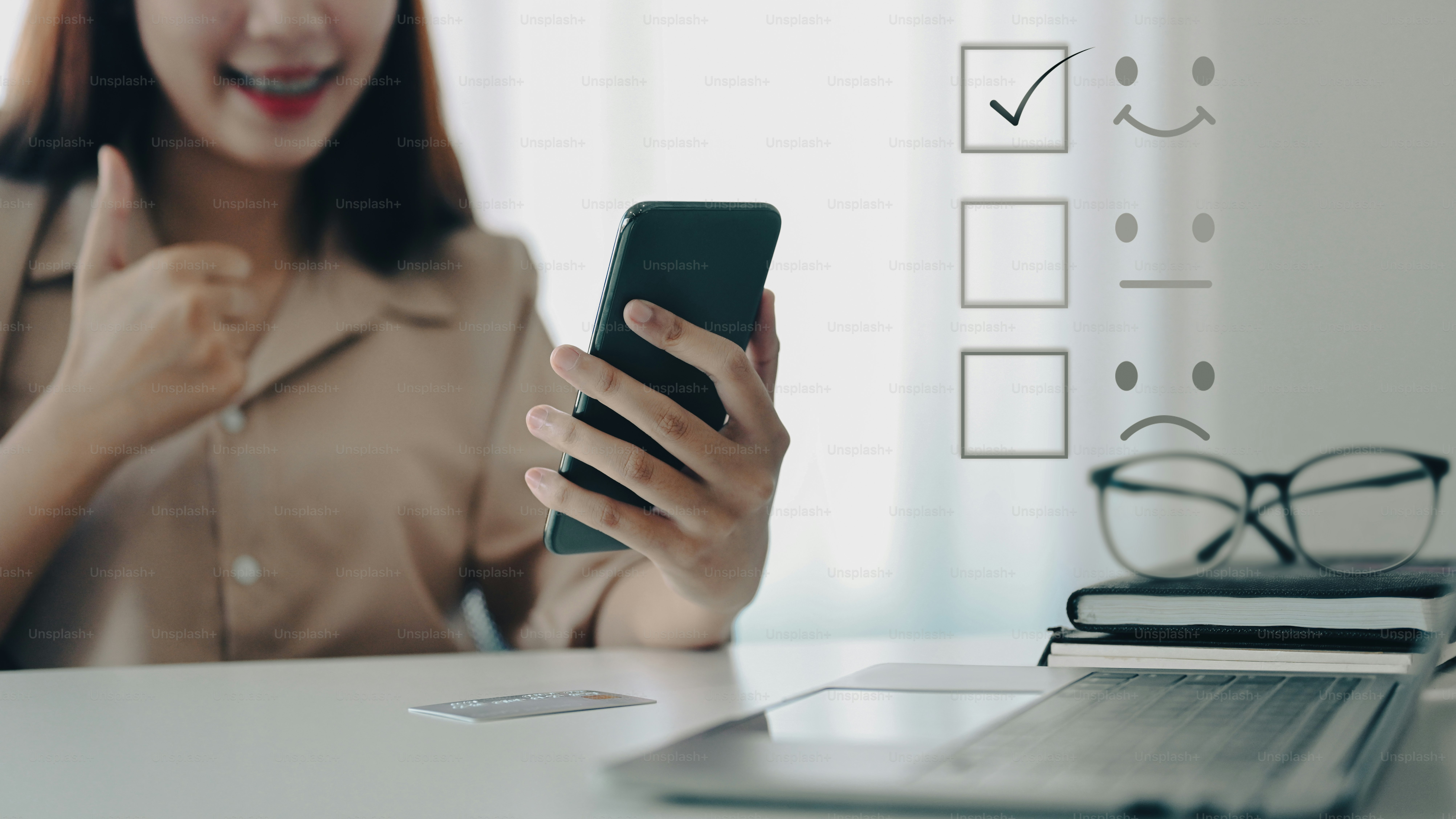 Smiling asian woman with thumb up and using smart phone for giving satisfied rating with smiley face icon to evaluate service.