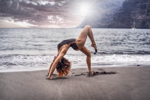 Attractive woman doing power pilates or hatha yoga on the beach with a beautiful view and moody sky. Healthy lifestyle. Silhouette of a girl in a yoga pose.