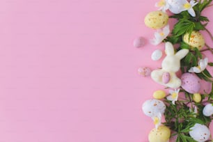 カラフルなイースターチョコレートの卵、ウサギ、春の花はピンクの背景にフラットに横たわっています。イースターおめでとう!スタイリッシュなイースターレイアウト。グリーティングカードまたはバナーテンプレート
