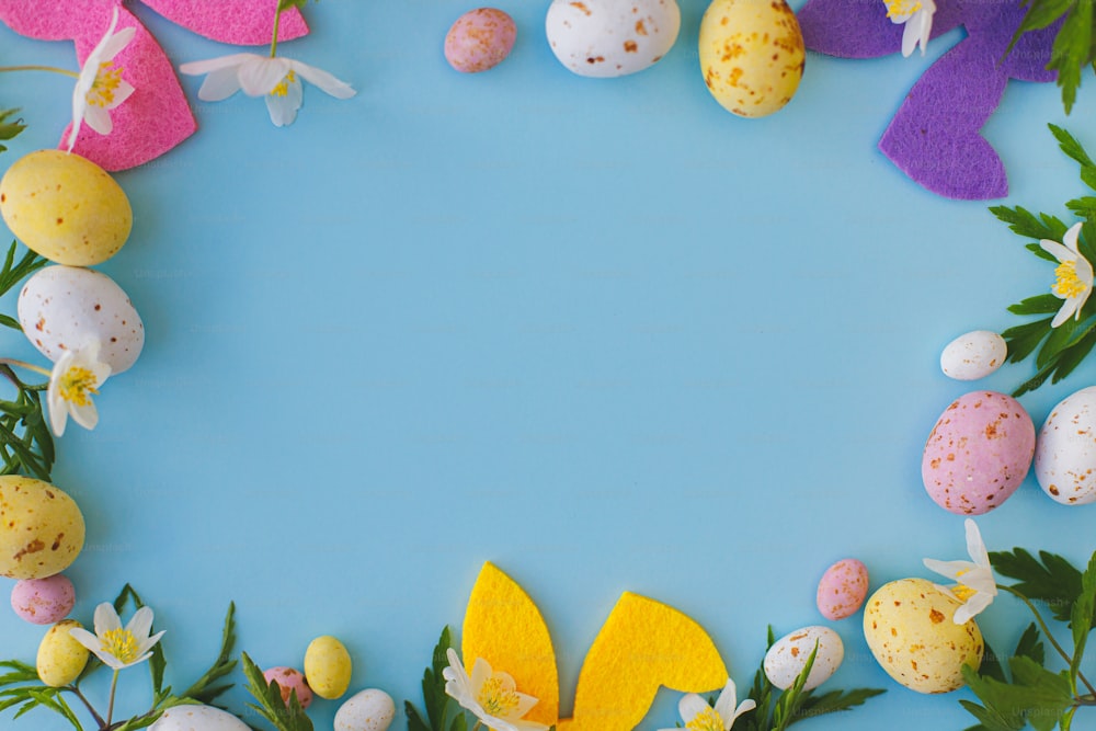 Colorful Easter bunnies, chocolate eggs and spring flowers frame on blue background, flat lay with space for text. Happy Easter! Pink and yellow artificial decor and fresh flowers. Easter hunt