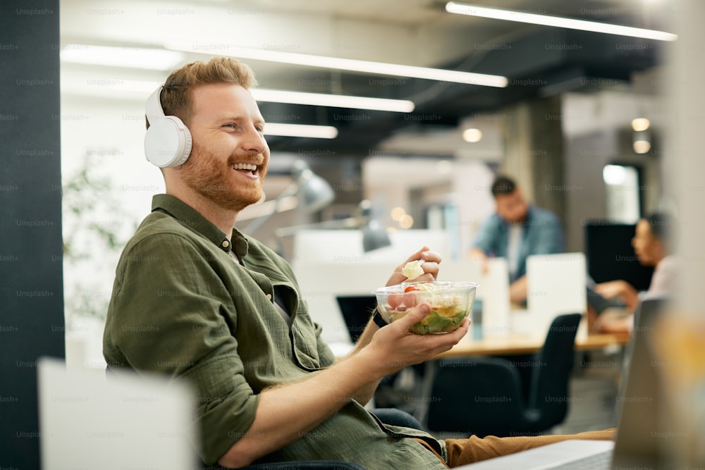 Feliz emprendedor disfrutando mientras almuerza y escucha música con auriculares en la oficina.