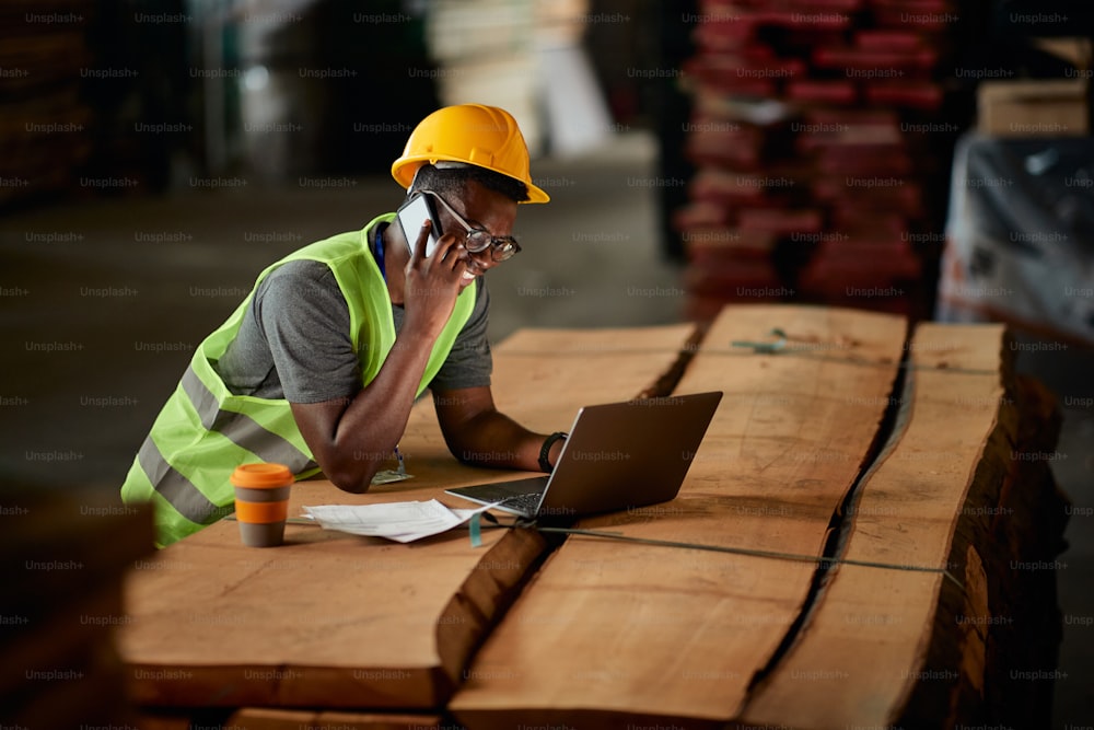 Happy African American worker using computer while communicating over cell phone at lumber warehouse.