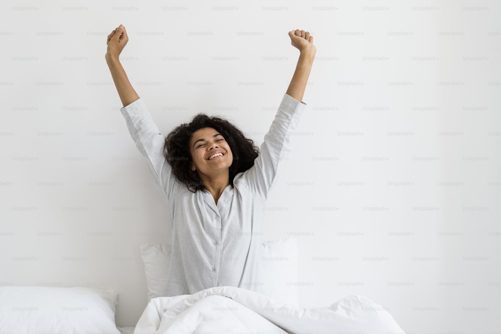 Beautiful female wearing in pajamas raised hands up, stretching body and smiling wide. Happy dark skinned woman waking up at home, spending early morning in bright cozy bedroom
