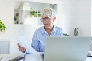 Focused old woman with white hair at home using laptop. Senior stylish entrepreneur working on computer at home. Woman analyzing and managing domestic bills and home finance