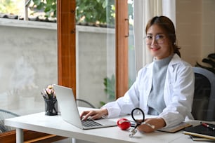 Doctora segura de sí misma en uniforme blanco sentada en el consultorio médico y sonriendo a la cámara. Concepto sanitario y médico.