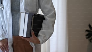 Cropped image of doctor in white uniform holding books and standing in medical office.