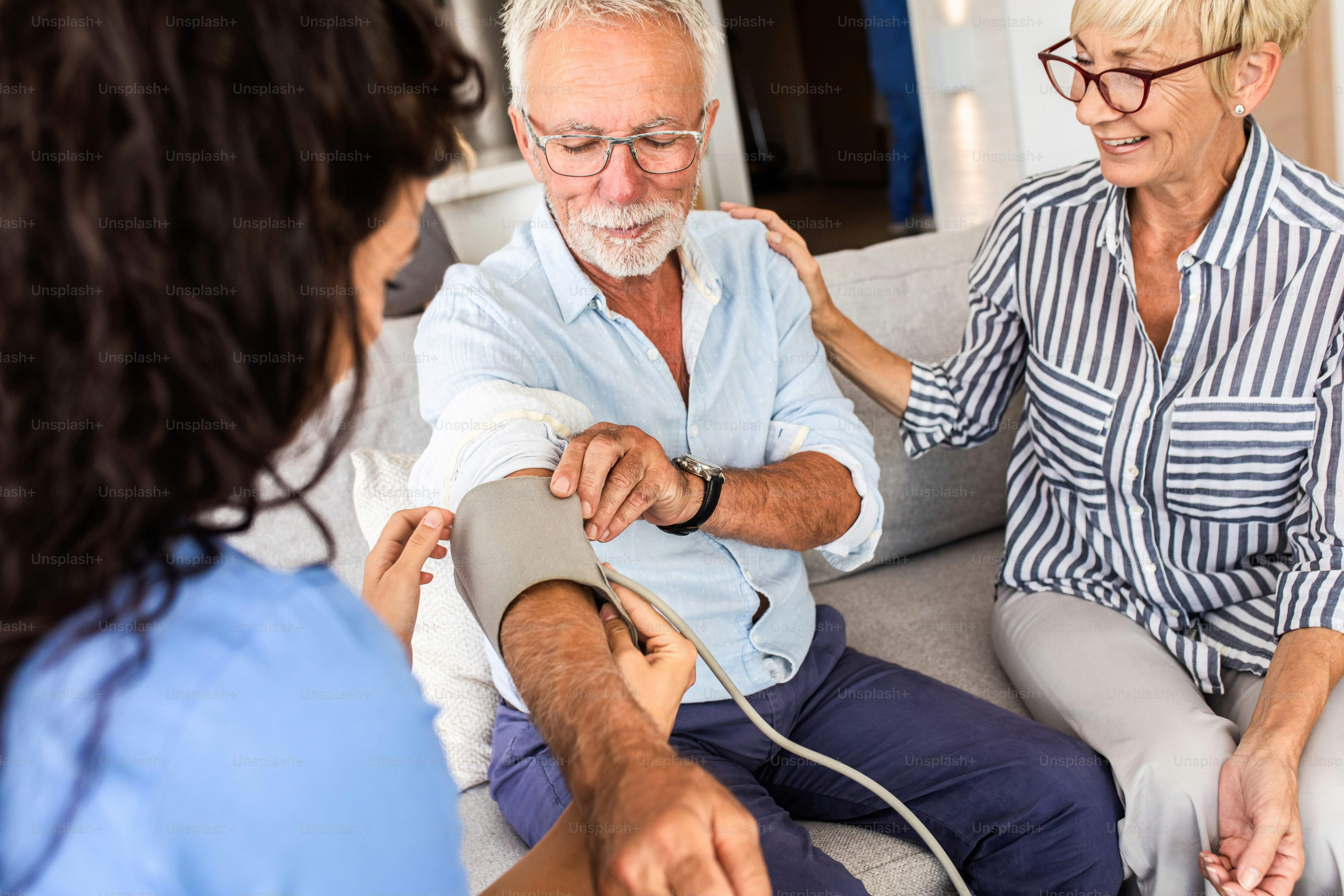 Female nurse talking to seniors patients while being in a home visit measuring blood pressure.