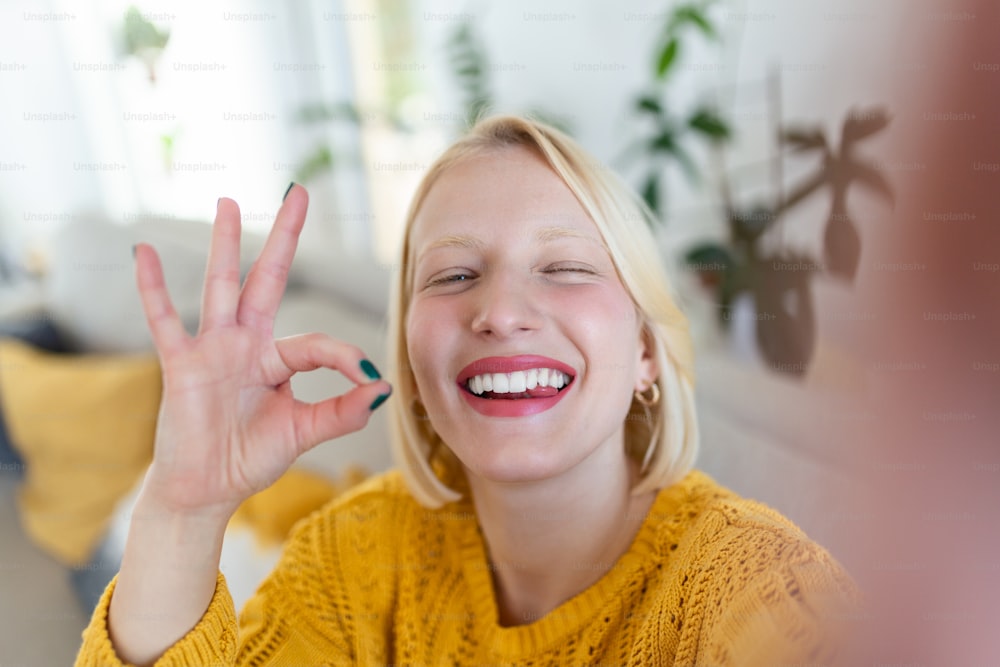 Schelmische junge Frau macht Selfie zu Hause. Schöne blonde Frau ok Schild mit Hand blinzelnd mit einem Auge und mit herausgestreckter Zunge