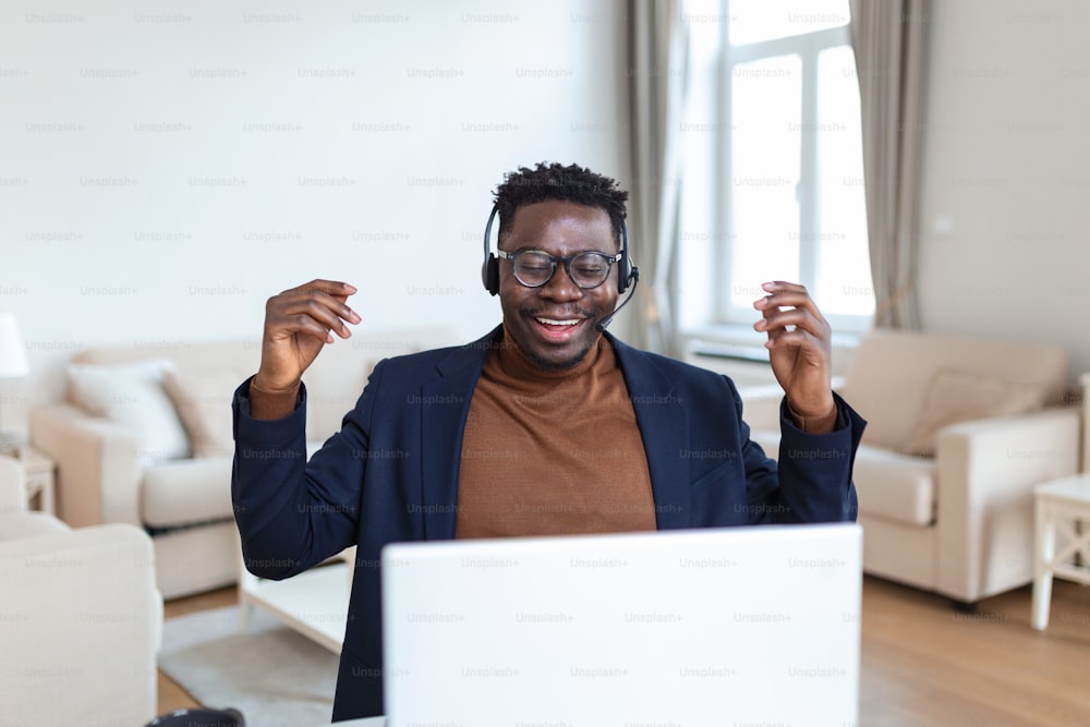 Aufgeregter afroamerikanischer Mann mit Kopfhörern, der gute Nachrichten in E-Mails liest, einen neuen Job bekommt, befördert wird, einen Laptop benutzt, auf den Bildschirm schaut und vor Freude schreit, Ja-Geste zeigt, feiert